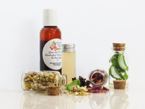 A left view of an all-natural facial toner in an amber bottle surrounded by small, corked glass bottles containing sprinkles of Red Rose, Aloe Vera and Chamomile ingredients. A clear glass bottle showcases the toner's color and texture.