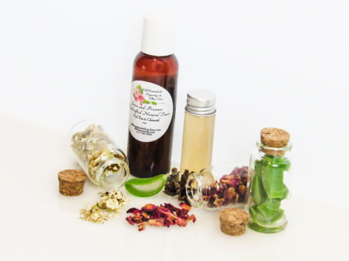 A right-angled frontal view of an all-natural facial toner in an amber bottle surrounded by small, corked glass bottles containing sprinkles of Red Rose, Aloe Vera and Chamomile ingredients. A clear glass bottle showcases the toner's color and texture.