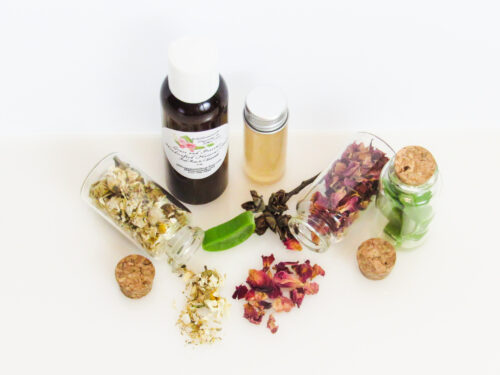 A top front view of an all-natural facial toner in an amber bottle surrounded by small, corked glass bottles containing sprinkles of Red Rose, Aloe Vera and Chamomile ingredients. A clear glass bottle showcases the toner's color and texture.