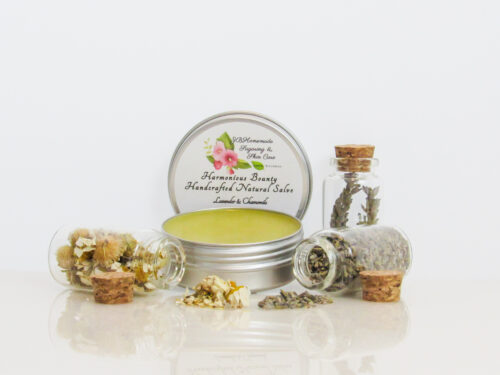 Harmonious Bounty presents its Creamy Natural Skin Salve in Lavender Chamomile, providing a comforting solution for dry skin. The product is showcased in an front view of an open tin, with its lid placed behind revealing the label. Encircling the salve are petite corked glass bottles filled with Lavender and Chamomile, accompanied by sprinkles of these soothing ingredients.