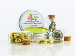 Harmonious Bounty presents its Creamy Natural Skin Salve in Lavender Chamomile, providing a comforting solution for dry skin. The product is showcased in a left-angled view of an open tin, with its lid placed behind revealing the label. Encircling the salve are petite corked glass bottles filled with Lavender and Chamomile, accompanied by sprinkles of these soothing ingredients.