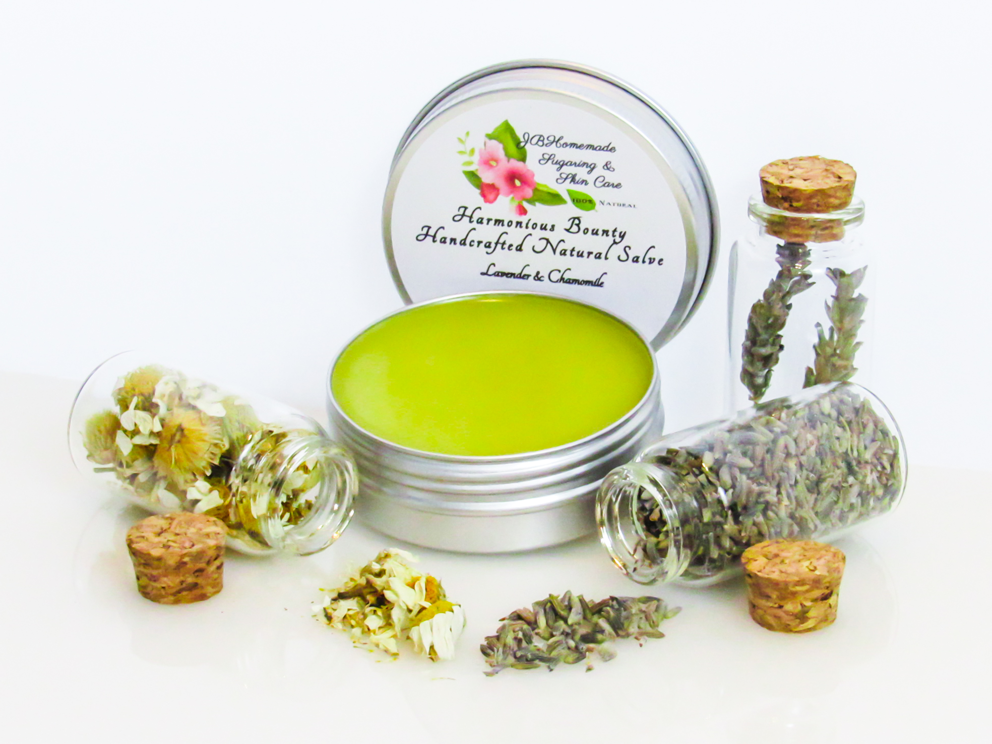 Harmonious Bounty presents its Creamy Natural Skin Salve in Lavender Chamomile, providing a comforting solution for dry skin. The product is showcased in an overhead right-angled view of an open tin, with its lid placed behind revealing the label. Encircling the salve are petite corked glass bottles filled with Lavender and Chamomile, accompanied by sprinkles of these soothing ingredients.