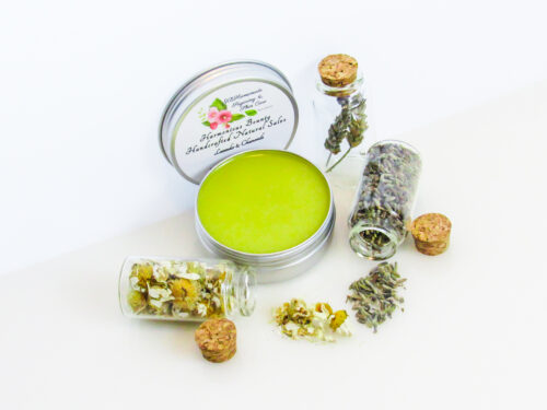 Harmonious Bounty presents its Creamy Natural Skin Salve in Lavender Chamomile, providing a comforting solution for dry skin. The product is showcased in an overhead left-angled view of an open tin, with its lid placed behind revealing the label. Encircling the salve are petite corked glass bottles filled with Lavender and Chamomile, accompanied by sprinkles of these soothing ingredients.