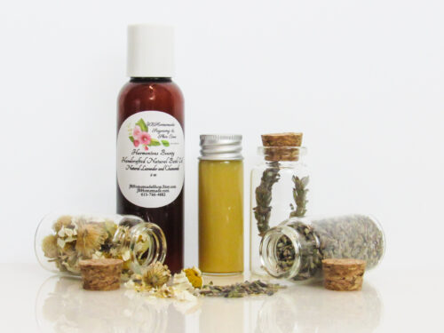 A front view of an all-natural bath oil in an amber bottle surrounded by small, corked glass bottles containing Lavender and Chamomile ingredients and sprinkles of the same. A clear glass bottle showcases the bath oil's color and texture.