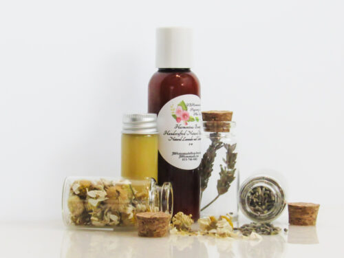 A left view of an all-natural bath oil in an amber bottle surrounded by small, corked glass bottles containing Lavender and Chamomile ingredients and sprinkles of the same. A clear glass bottle showcases the bath oil's color and texture.