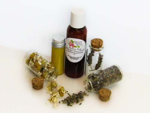 A slightly angled frontal view of an all-natural bath oil in an amber bottle surrounded by small, corked glass bottles containing Lavender and Chamomile ingredients and sprinkles of the same. A clear glass bottle showcases the bath oil's color and texture.