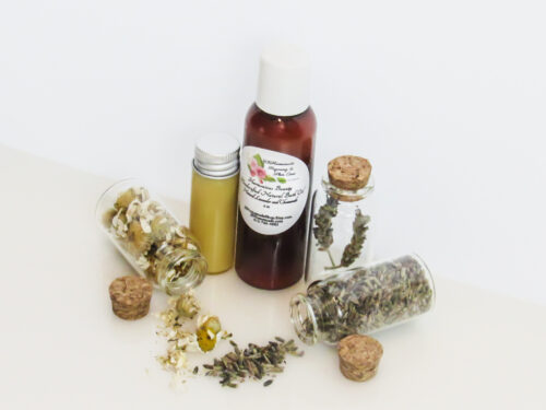 An angled, front view of an all-natural bath oil in an amber bottle surrounded by small, corked glass bottles containing Lavender and Chamomile ingredients and sprinkles of the same. A clear glass bottle showcases the bath oil's color and texture.