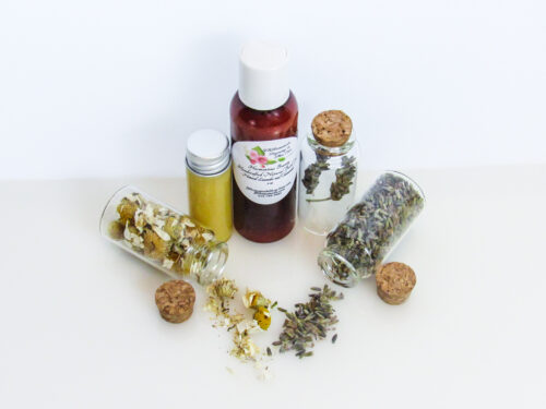 An angled, top view of an all-natural bath oil in an amber bottle surrounded by small, corked glass bottles containing Lavender and Chamomile ingredients and sprinkles of the same. A clear glass bottle showcases the bath oil's color and texture.