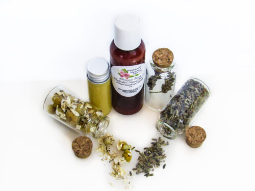A top angled view of an all-natural bath oil in an amber bottle surrounded by small, corked glass bottles containing Lavender and Chamomile ingredients and sprinkles of the same. A clear glass bottle showcases the bath oil's color and texture.