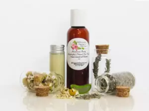 A front view of an all-natural facial cleanser in an amber bottle surrounded by small, corked glass bottles containing Lavender, Aloe Vera and Chamomile ingredients and sprinkles of the same. A clear glass bottle showcases the face wash's color and texture.