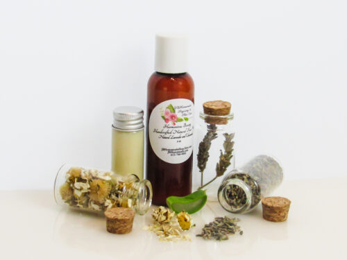 A front view of an all-natural facial cleanser in an amber bottle surrounded by small, corked glass bottles containing Lavender, Aloe Vera and Chamomile ingredients and sprinkles of the same. A clear glass bottle showcases the face wash's color and texture.