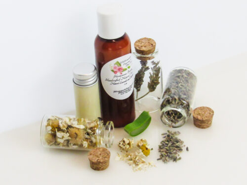 An angled, left view of an all-natural facial cleanser in an amber bottle surrounded by small, corked glass bottles containing Lavender, Aloe Vera and Chamomile ingredients and sprinkles of the same. A clear glass bottle showcases the face wash's color and texture.