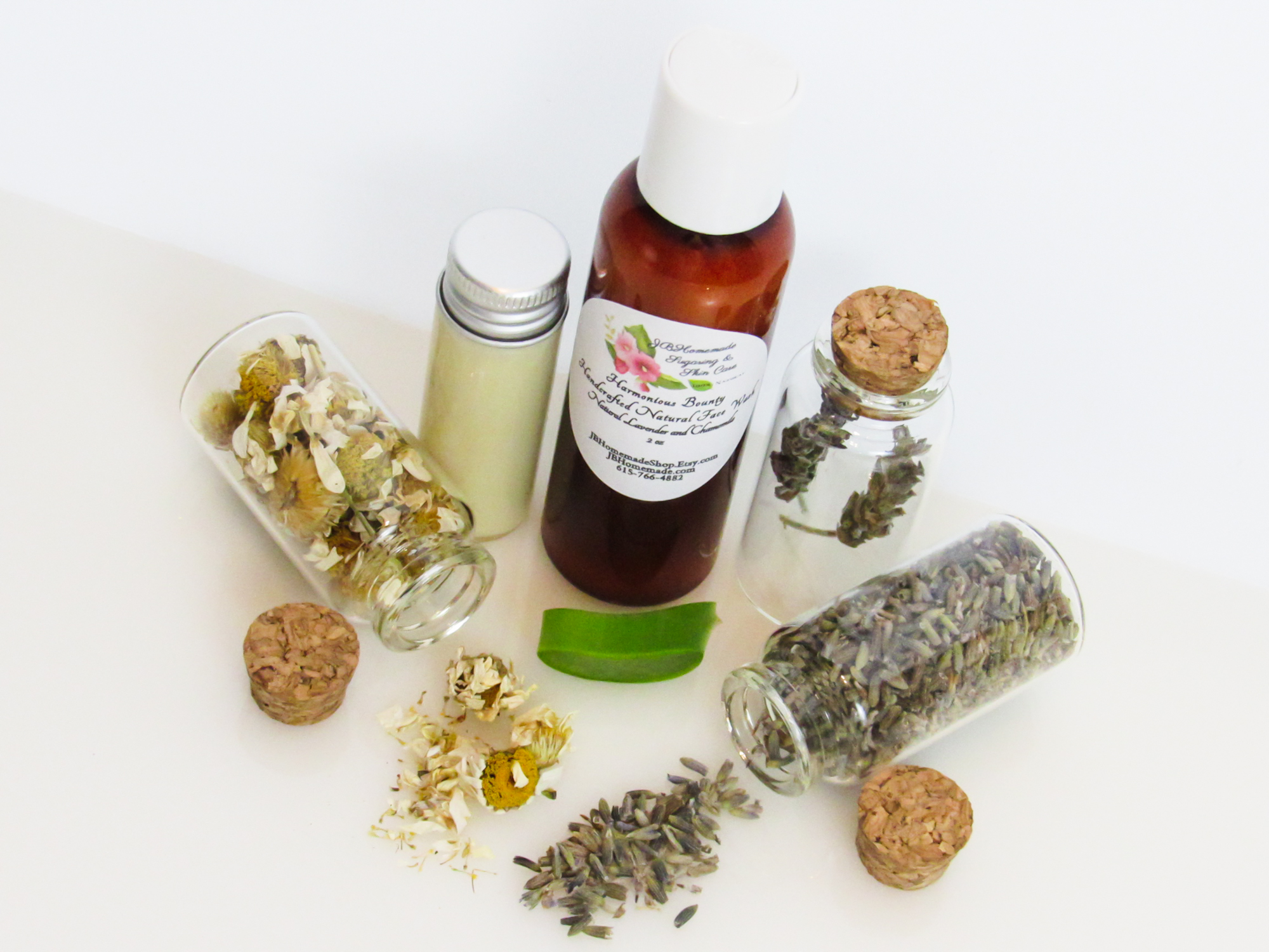 An angled, top view of an all-natural facial cleanser in an amber bottle surrounded by small, corked glass bottles containing Lavender, Aloe Vera and Chamomile ingredients and sprinkles of the same. A clear glass bottle showcases the face wash's color and texture.
