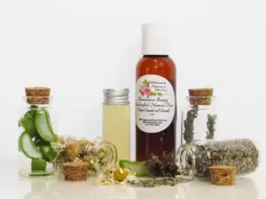 A front view of an all-natural toner in an amber bottle surrounded by small, corked glass bottles containing sprinkles of Lavender, Aloe Vera and Chamomile ingredients and sprinkles of the same. A clear glass bottle showcases the toner's color and texture.