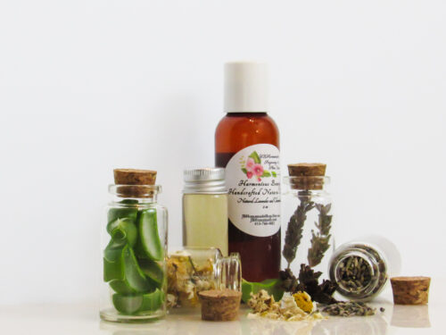 A left front view of an all-natural toner in an amber bottle surrounded by small, corked glass bottles containing sprinkles of Lavender, Aloe Vera and Chamomile ingredients and sprinkles of the same. A clear glass bottle showcases the toner's color and texture.