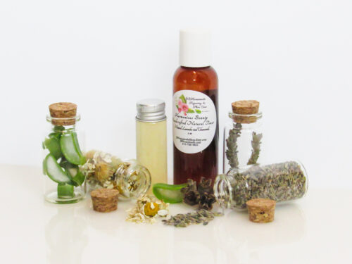A front view of an all-natural toner in an amber bottle surrounded by small, corked glass bottles containing sprinkles of Lavender, Aloe Vera and Chamomile ingredients and sprinkles of the same. A clear glass bottle showcases the toner's color and texture.