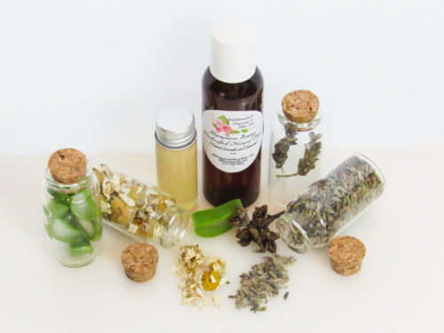 A front view of an all-natural toner in an amber bottle surrounded by small, corked glass bottles containing sprinkles of Lavender, Aloe Vera and Chamomile ingredients and sprinkles of the same. A clear glass bottle showcases the toner's color and texture.