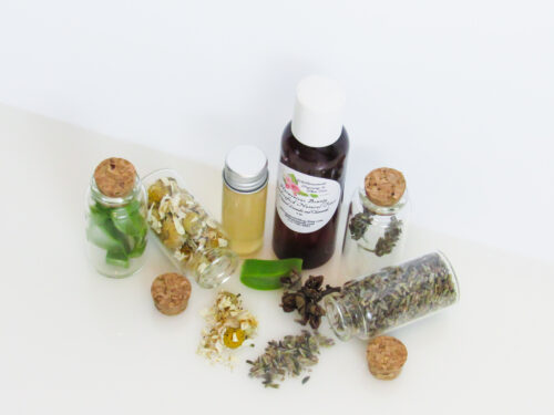 A front top angled view of an all-natural toner in an amber bottle surrounded by small, corked glass bottles containing sprinkles of Lavender, Aloe Vera and Chamomile ingredients and sprinkles of the same. A clear glass bottle showcases the toner's color and texture.