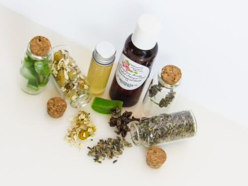 A top right-angled view of an all-natural toner in an amber bottle surrounded by small, corked glass bottles containing sprinkles of Lavender, Aloe Vera and Chamomile ingredients and sprinkles of the same. A clear glass bottle showcases the toner's color and texture.