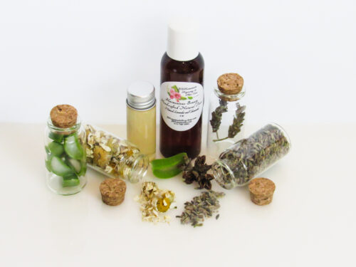 A front angled view of an all-natural toner in an amber bottle surrounded by small, corked glass bottles containing sprinkles of Lavender, Aloe Vera and Chamomile ingredients and sprinkles of the same. A clear glass bottle showcases the toner's color and texture.
