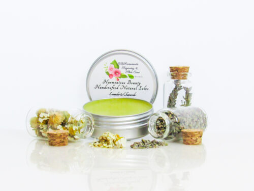 Harmonious Bounty presents its Creamy Natural Skin Salve in Lavender Chamomile, providing a comforting solution for dry skin. The product is showcased in a front view of an open tin, with its lid placed behind revealing the label. Encircling the salve are petite corked glass bottles filled with Lavender and Chamomile, accompanied by sprinkles of these soothing ingredients.