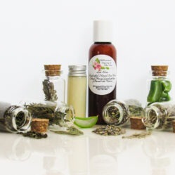 Front perspective view of an all-natural facial cleanser in an amber bottle surrounded by six small, corked glass bottles containing sprinkles of pine, cedarwood, spearmint, and lavender ingredients. A clear glass bottle showcases the face wash’s color and texture.