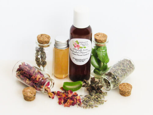 Naturally Serene Lavender and Red Rose Toner in an amber bottle, surrounded by organic ingredients like aloe vera slices, dried lavender sprigs, witch hazel, and red rose petals on a white surface.