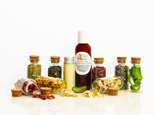 An all-natural facial cleanser in an amber bottle surrounded by eight small corked glass bottles containing sprinkles of Calendula, Rose, Lavender, Chamomile, Rosemary, Aloe Vera, Sandalwood and Patchouli ingredients and sprinkles of the same. A clear glass bottle showcases the face wash's color and texture. Front view.