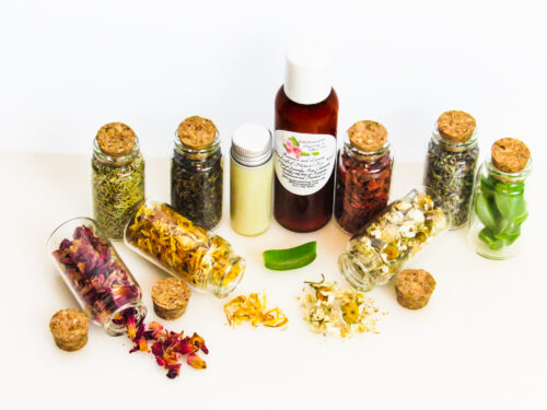 An all-natural facial cleanser in an amber bottle surrounded by eight small corked glass bottles containing sprinkles of Calendula, Rose, Lavender, Chamomile, Rosemary, Aloe Vera, Sandalwood and Patchouli ingredients and sprinkles of the same. A clear glass bottle showcases the face wash's color and texture. Overhead view.