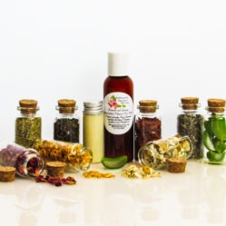 An all-natural facial cleanser in an amber bottle surrounded by eight small corked glass bottles containing sprinkles of Calendula, Rose, Lavender, Chamomile, Rosemary, Aloe Vera, Sandalwood and Patchouli ingredients and sprinkles of the same. A clear glass bottle showcases the face wash's color and texture. Front view.