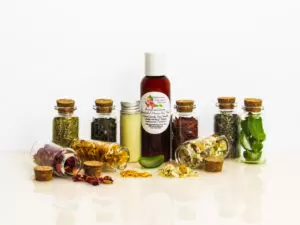 An all-natural facial cleanser in an amber bottle surrounded by eight small corked glass bottles containing sprinkles of Calendula, Rose, Lavender, Chamomile, Rosemary, Aloe Vera, Sandalwood and Patchouli ingredients and sprinkles of the same. A clear glass bottle showcases the face wash's color and texture. Front view.