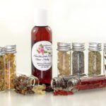 A bottle of Adorn Your Garden Sugaring Cleanser and Prep surrounded by small jars containing its all-natural ingredients like Chamomile, Calendula, Frankincense, Clove, Sandalwood, Lavender, Rosemary, and Ylang Ylang Extra. Front view.