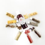 A bottle of Adorn Your Garden Sugaring Cleanser and Prep surrounded by small jars containing its all-natural ingredients like Chamomile, Calendula, Frankincense, Clove, Sandalwood, Lavender, Rosemary, and Ylang Ylang Extra. Overhead view.