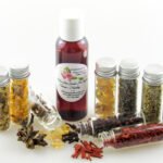 A bottle of Adorn Your Garden Sugaring Cleanser and Prep surrounded by small jars containing its all-natural ingredients like Chamomile, Calendula, Frankincense, Clove, Sandalwood, Lavender, Rosemary, and Ylang Ylang Extra. Front view.