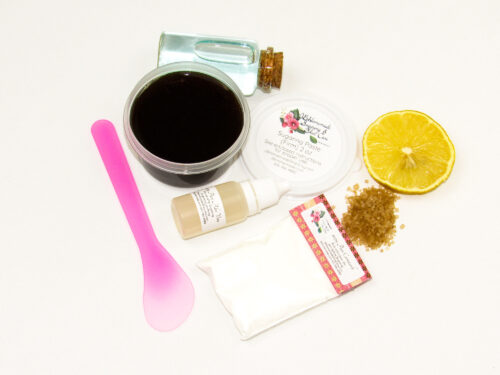 A 2-ounce tub of JBHomemade Sugaring Paste is presented with its included pouch of cornstarch, bottle of aloe vera and applicator next to a slice of fresh lemon, a glass jar filled with clear blue water, and another jar tipped over, spilling raw sugar, accentuating the natural ingredients.